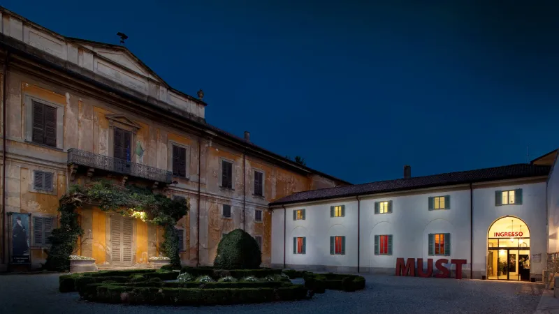 La corte di Villa Sottocasa al tramonto: la facciata ocra con il balcone e le bandiere, il parterre di siepi e l’ala bianca del MUST con l’ingresso illuminato e le grandi lettere rosse MUST