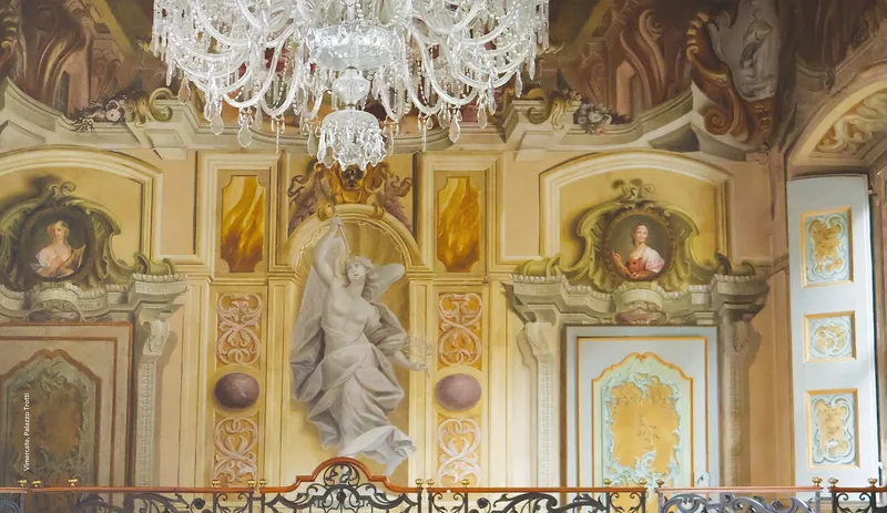 Una sala affrescata di Palazzo Trotti: quadrature barocche e rococò, figura alata in grisaille con la tromba, medaglioni con ritratti femminili e lampadario di cristallo
